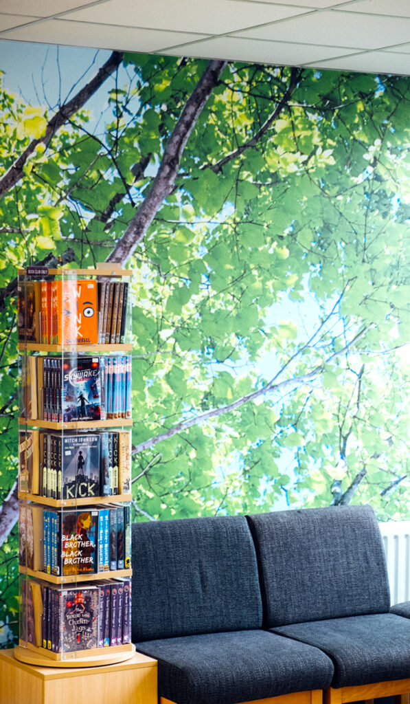 Wall art depicting tree branches heavily laden with leaves against blue sky background.  A revolving bookcase and seating are placed in front.