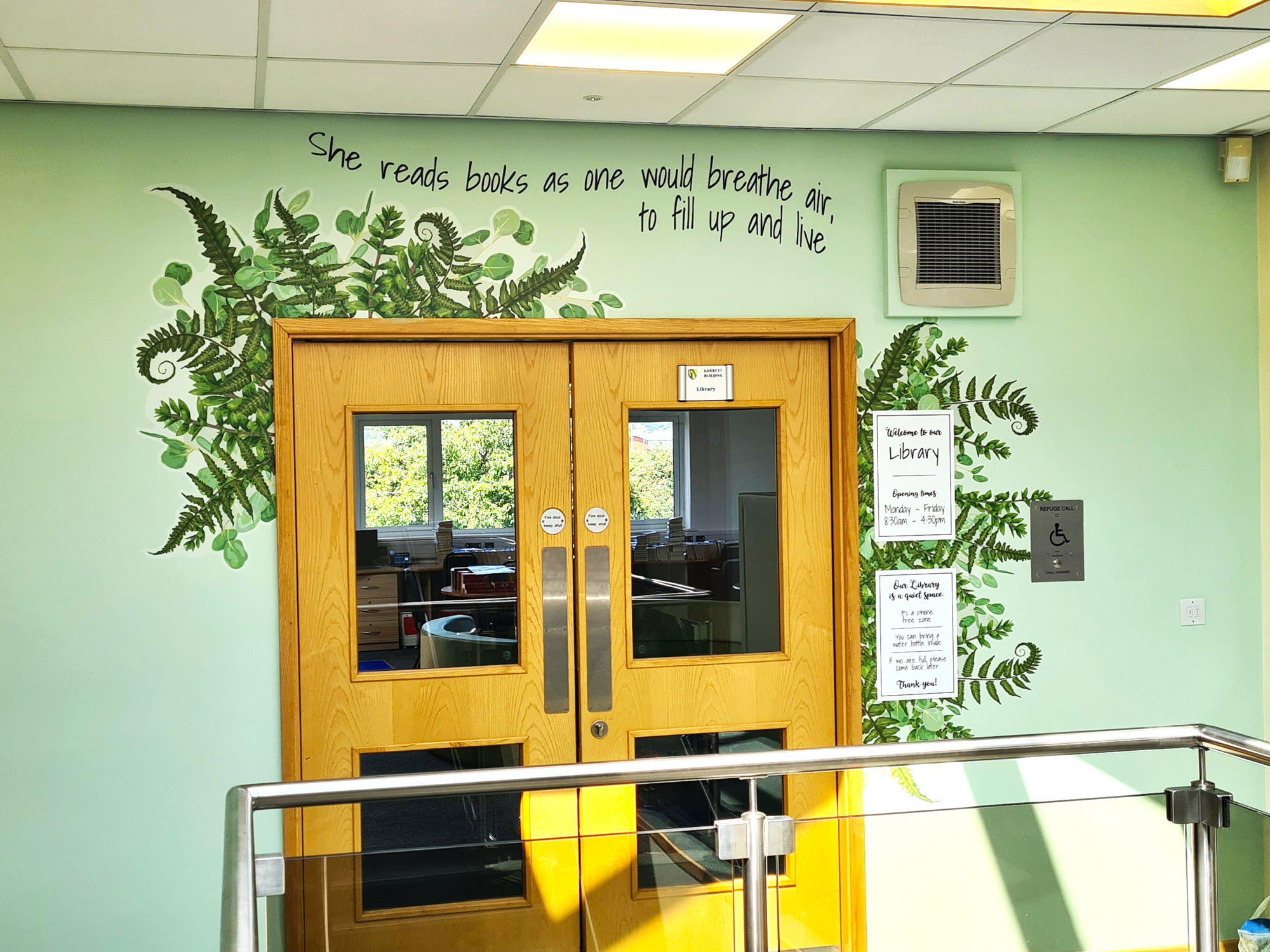 Mural of fern is shown on pale green background around the top edge of library entrance door with wording above the fern which reads "She reads books as one would breathe or, to fill up and live"