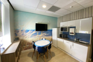 A room with a dining table and four blue chairs with wooden legs. White kitchen cupboards are separated by a blue wall and a wood effect mural is featured above the cabinets. The back wall features a mural of a seaside scene behind a wall mounted TV.