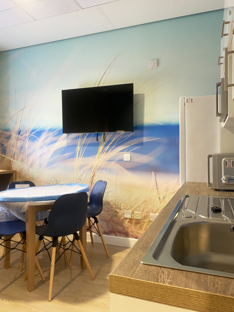 A room with a table and dining chairs with kitchen cabinets on one side.  The rear wall features a TV on top of wall art depicting soft focus seaside grasses.
