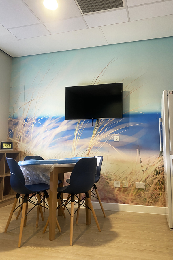 A room with a table and dining chairs with kitchen cabinets on one side.  The rear wall features a TV on top of wall art depicting soft focus seaside grasses.