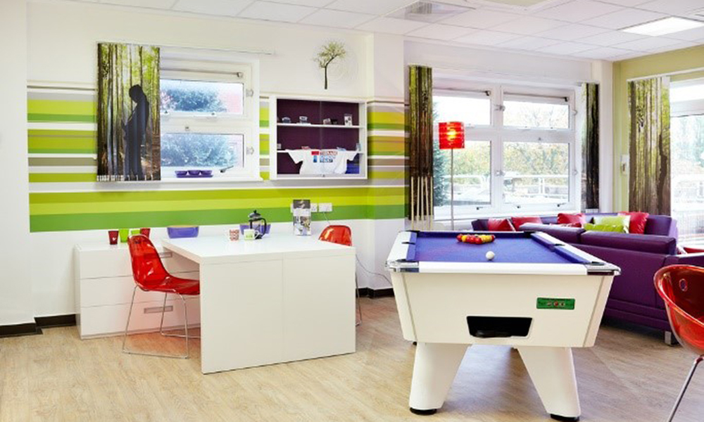 Teenage Cancer Trust social zone decorated in purple and green with striped wall art, pool table, table and chairs and sofas.
