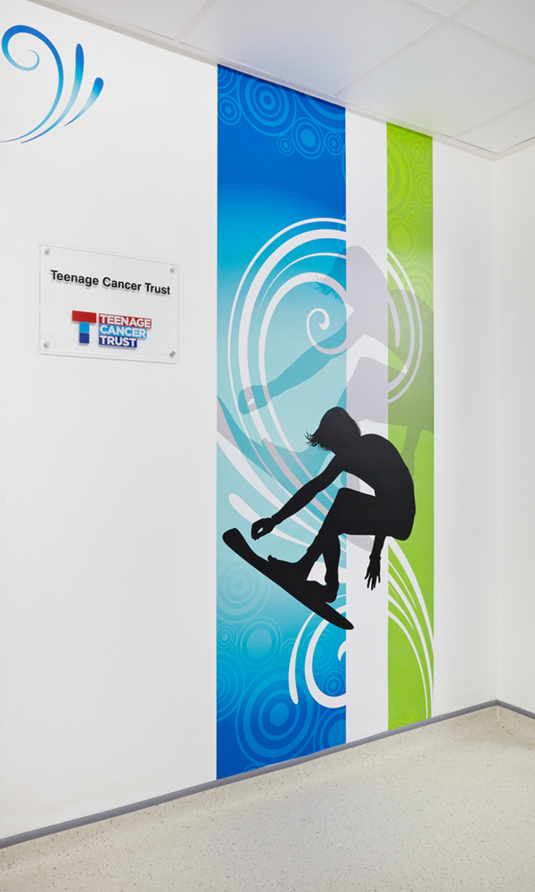 A wall with a Teenage Cancer Trust logo  and a silhouette of a person on a surfboard with blue and green swirls in the background.